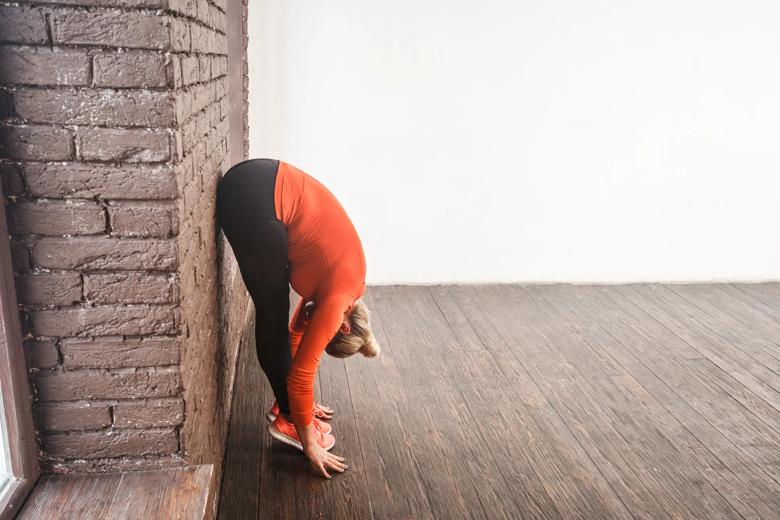 Leaning against a wall, a person bends down to the floor until their hands are touching it, standing on tiptoe