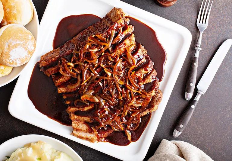 A plate of brisket topped with cooked onions with a fork and knife nearby