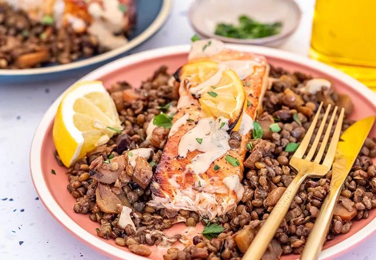 A plate with cooked salmon over lentils with mustard vinaigrette and slices of lemon.