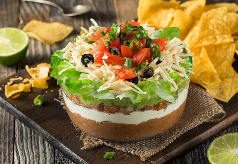 A bowl of eight-layer Mexican bean dip on a wooden table with chips in background.