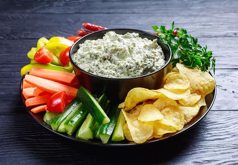 A bowl of artichoke dip surrounded by sliced carrots, peppers, cucumbers and potato chips
