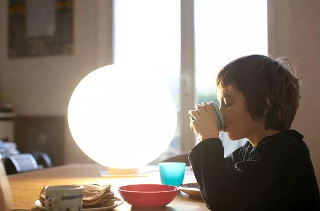 Child sitting at a table next to a lamp, eating and drinking.