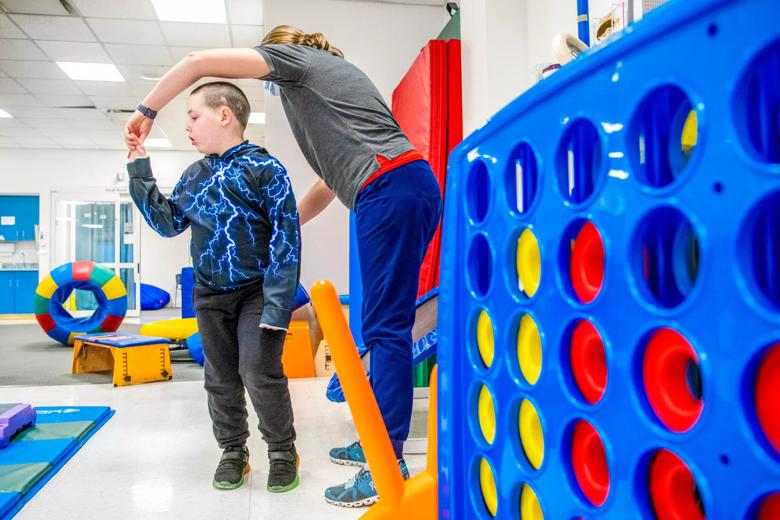 A caregiver helps a child turn around in a room filled with therapy toys