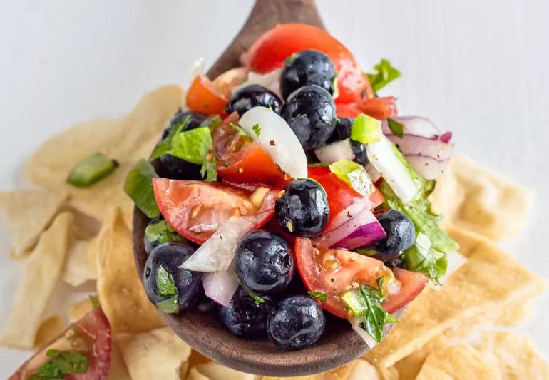 Closeup of blueberry jalapeno salsa in wooden spoon suspended over tortilla chips.