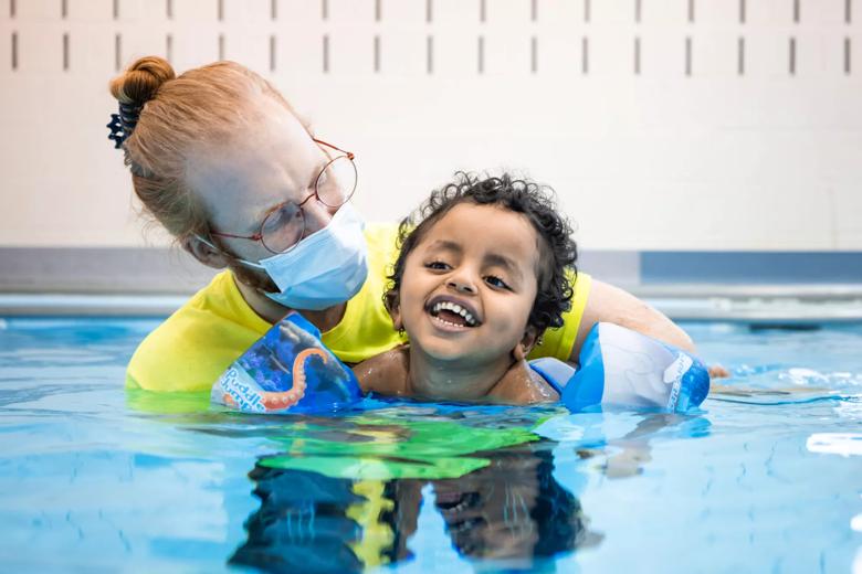 Person in a yellow shirt helps a child float in a pool