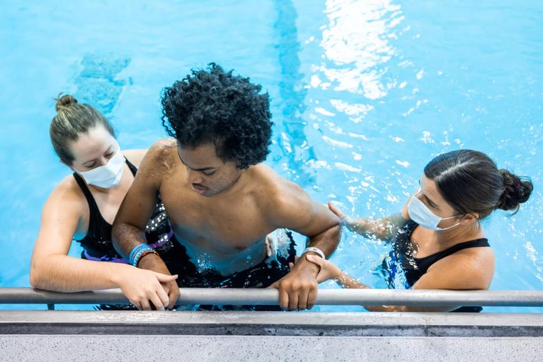 2 people help a person climb a ladder to get out of a pool