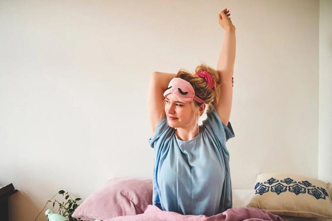 Person waking up, sitting up in bed, stretching, with sleep mask raised to forehead