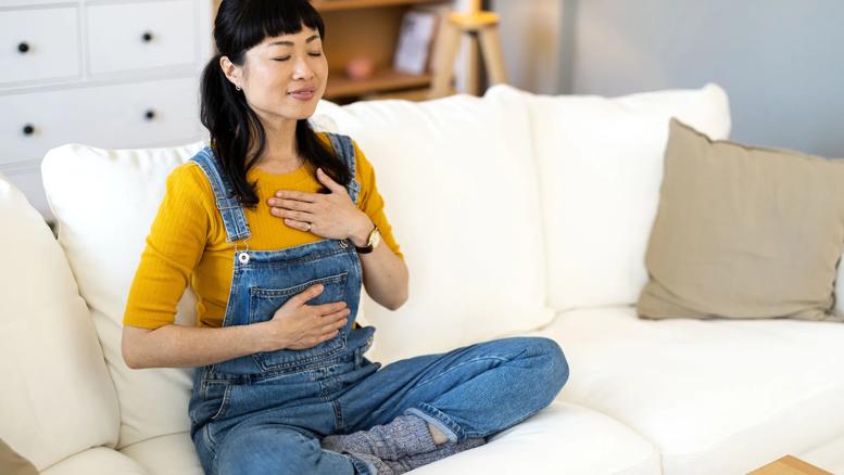 Woman sitting cross-legged on couch, hand on abdomen and hand on chest, meditating