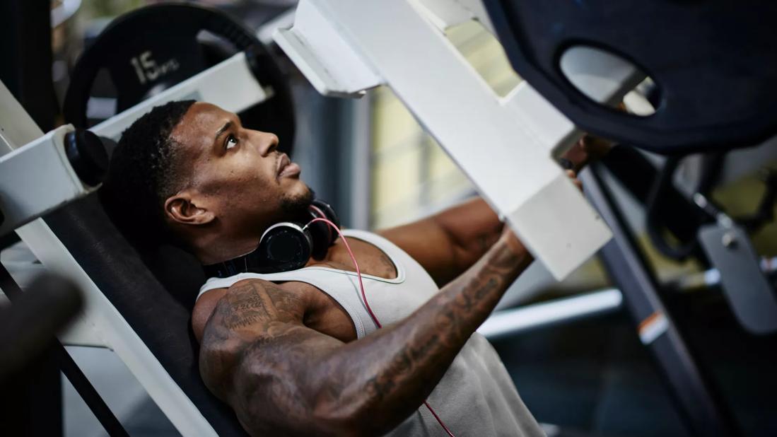 Muscular person using weight machine in gym, headphones around neck