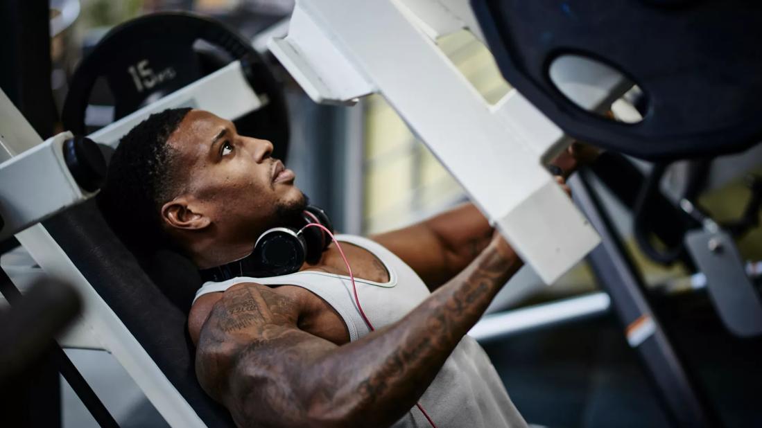 Muscular person using weight machine in gym, headphones around neck