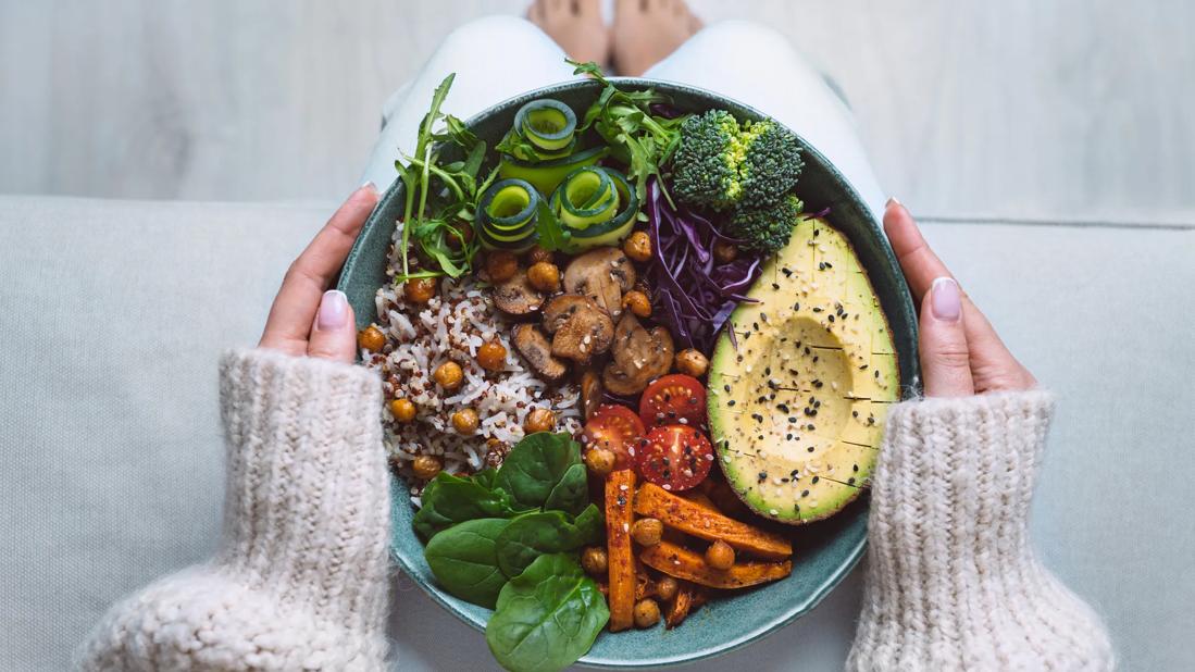 Person holding bowl of vegetarian foods on their lap, like avocado, rice, veggies and chickpeas