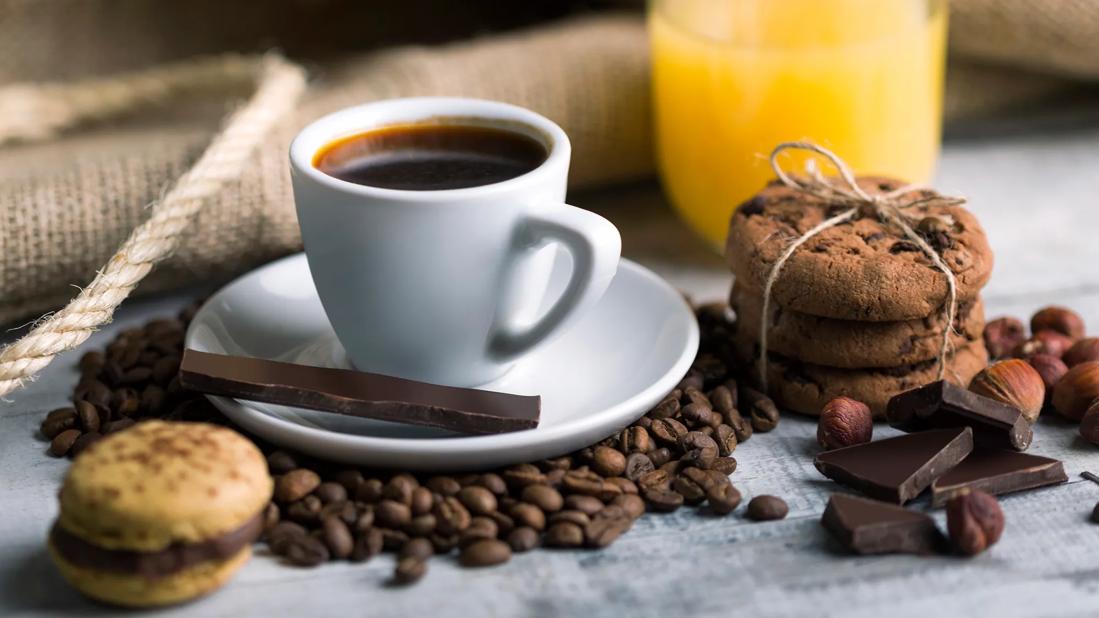 Cup of coffee on saucer, with scattered coffee beans and chocolate chunks, and baked goods