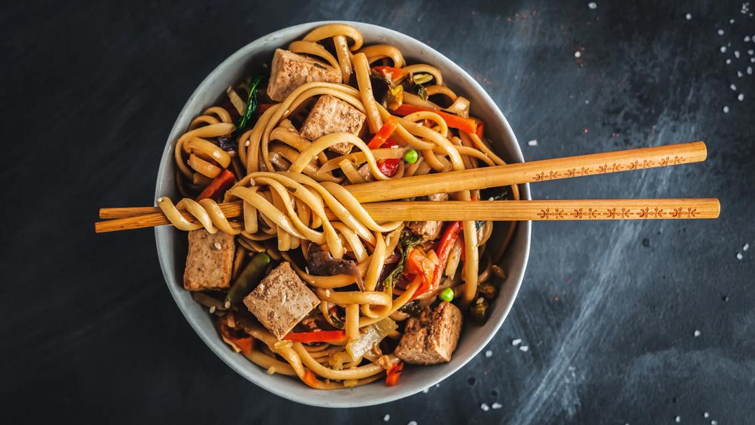 Bowl of soba noodles with veggies and tofu, with chopsticks across top of bowl