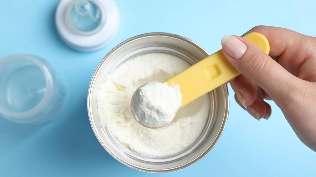 Close up of hand holding a scoop of powder baby formula over container of powder baby formula