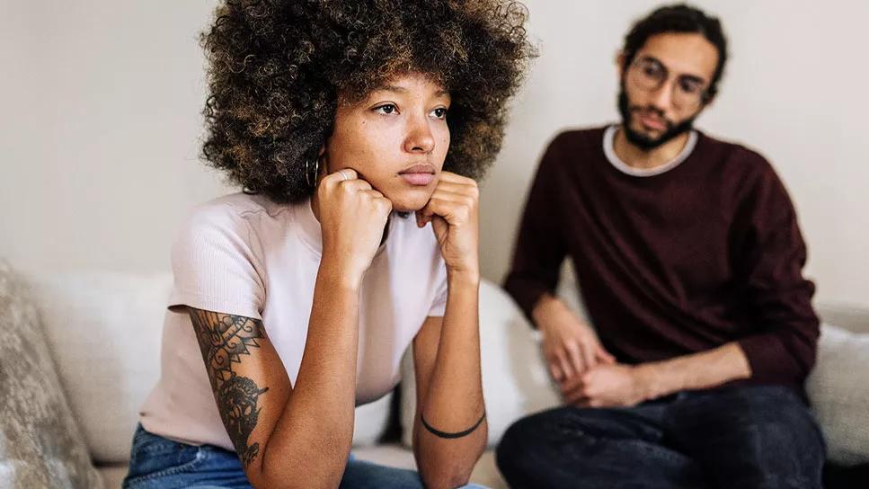 Female sitting on couch staring blankly ahead, with male next to her on couch staring at her