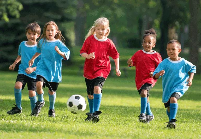 Children on two different soccer teams running after soccer ball in field.