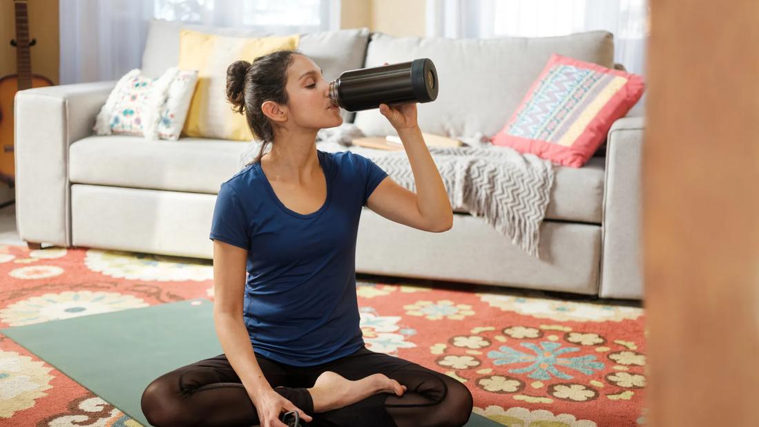 Person sitting on yoga mat in living room, drinking from bottle of water, cell phone on mat