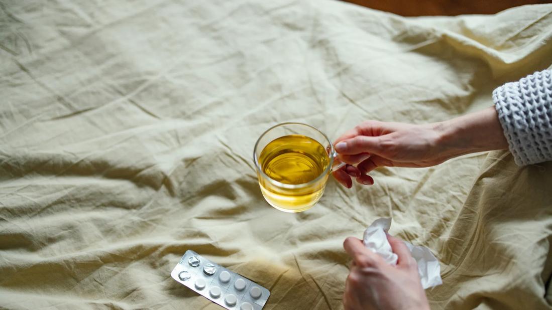 Hand holding hot glass mug of tea, with medicine packet open on bed cover