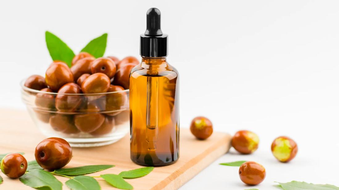 Glass bottle of jojoba oil, next to jojoba fruits in a bowl and scattered fruits and leaves