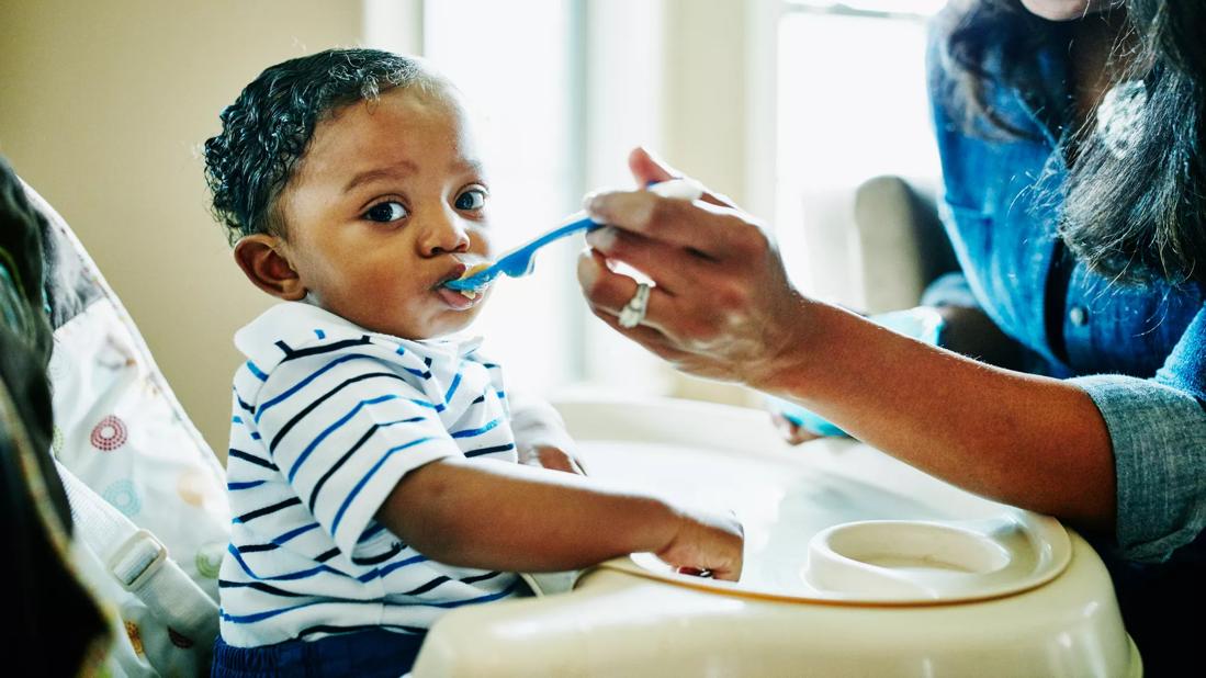 A toddler in a highchair wearing a striped shirt eats food off a spoon held by a parent