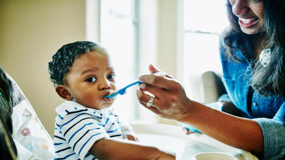 A toddler in a highchair wearing a striped shirt eats food off a spoon held by a parent