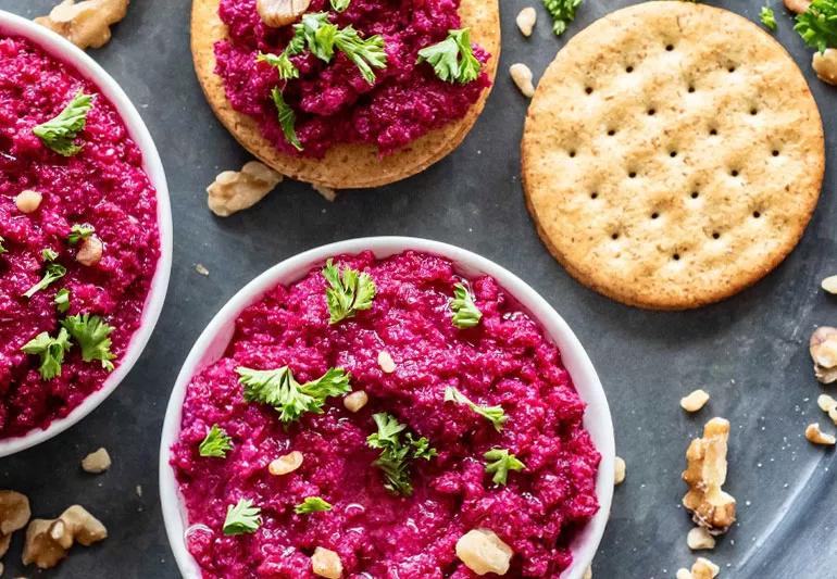 Aerial view of bright purple beet muhammara in small white bowls and atop crackers