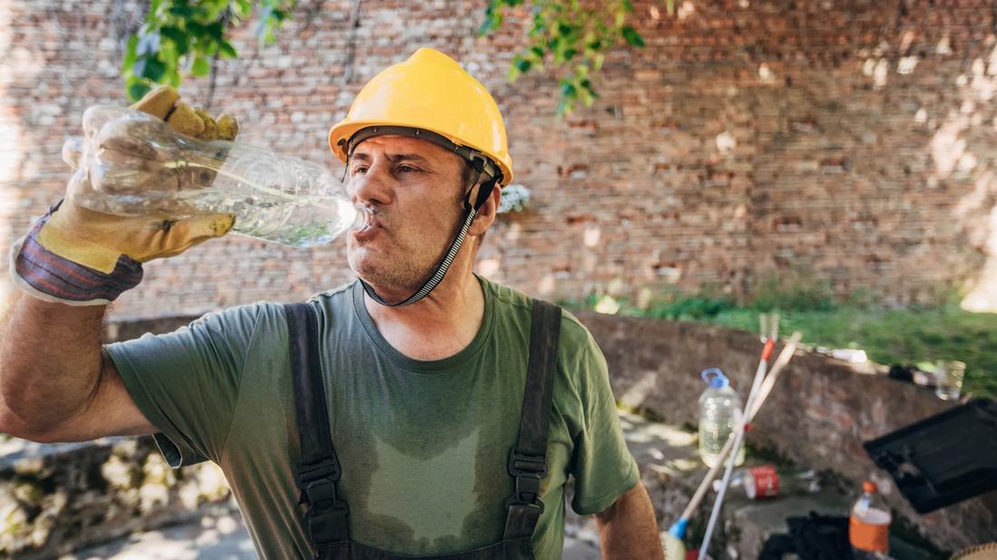 Sweaty construction worker in yellow hard hat drinking water from a plastic bottle