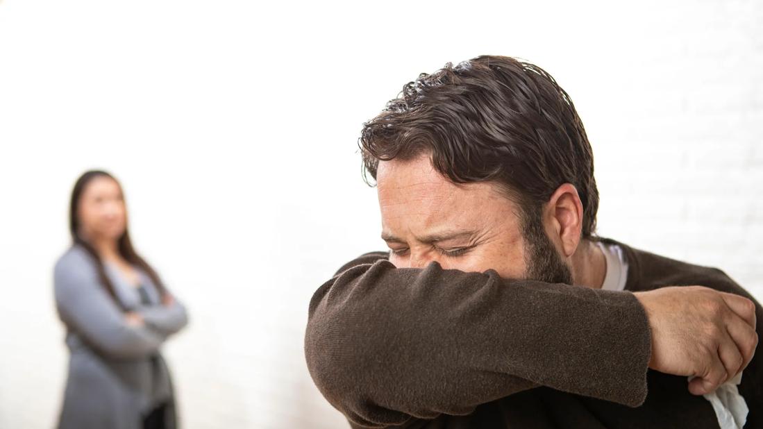 Person sneezing into arm, holding tissue, blurry person in background