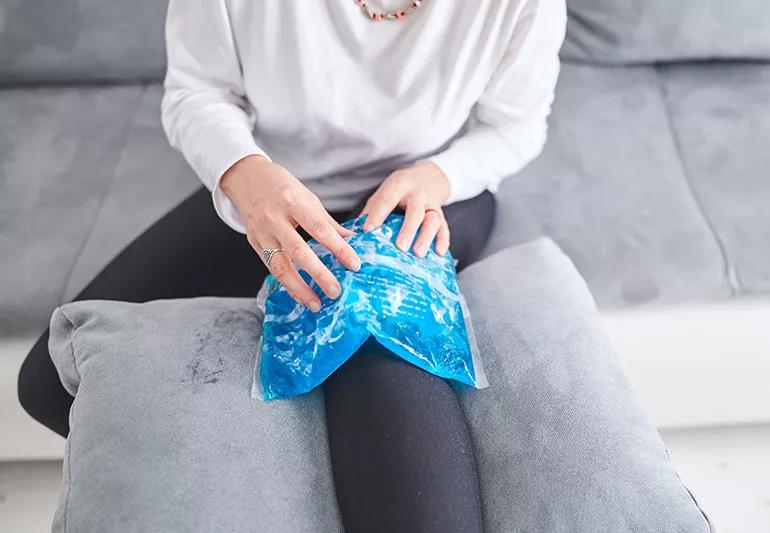 A person sits on the couch with their leg elevated while they place an ice pack on their knee