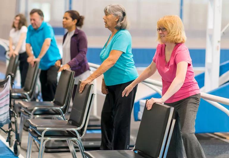 Group of elderly working on balancing with chairs for support