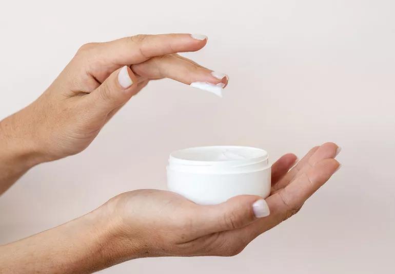 A person dips their fingers into a small topical ointment container.