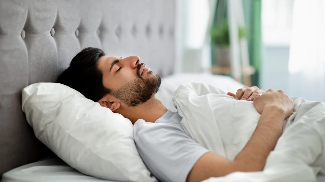 Person lying on back, hands folded against chest, sleeping in bright daylit room