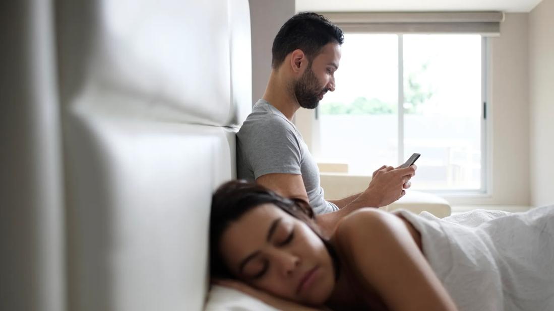 A couple in bed, one sleeping, one on their cellphone, in early morning light