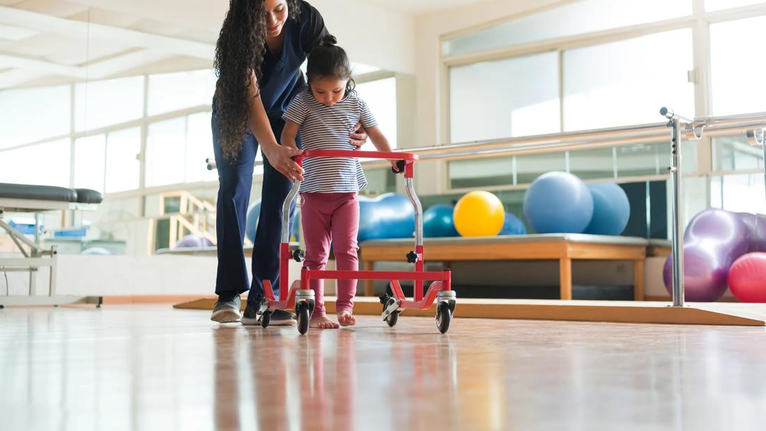 Healthcare provider helping child walk with a walker in therapy room