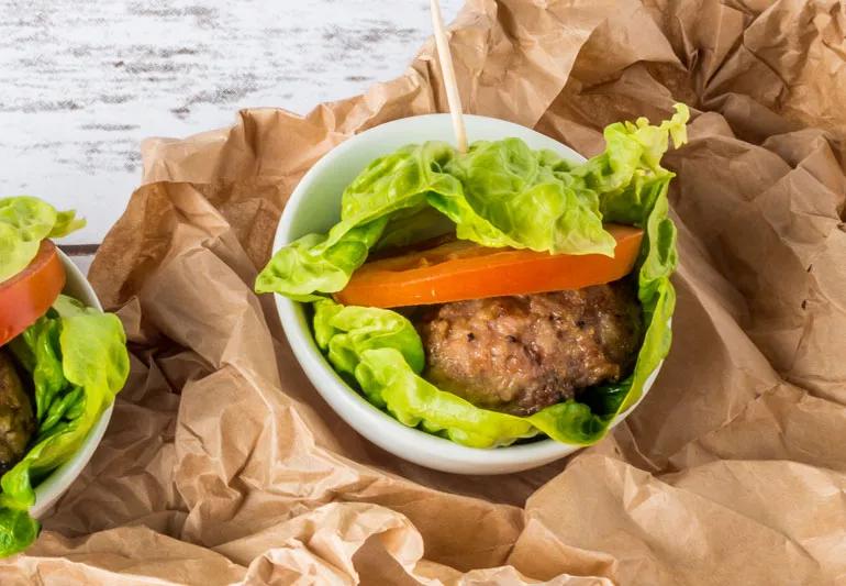 Burger and tomato wrapped in lettuce, sitting in a white bowl atop a crumpled brown bag