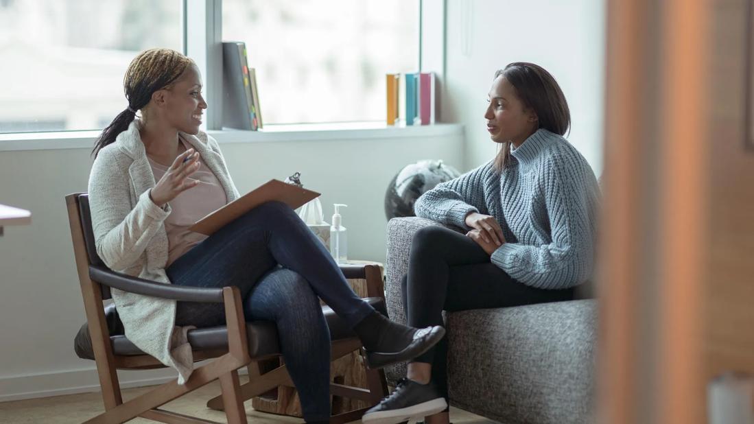 A therapist sitting in chair holding a clipboard and a patient sitting on couch talking in office