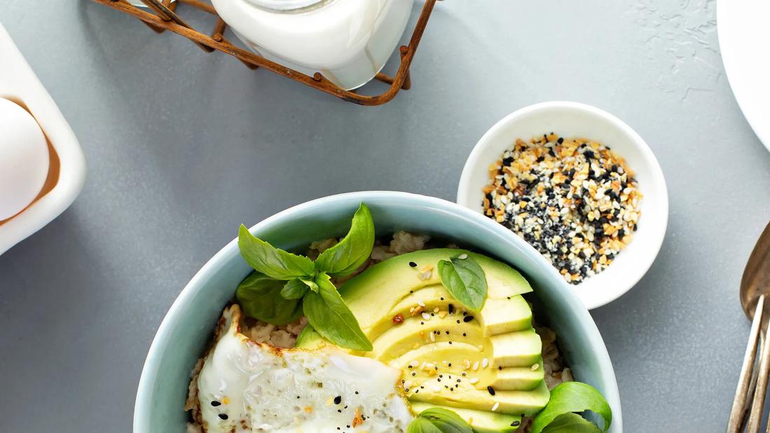 Bowl of oatmeal, topped with fried egg and avocado, with small bowl of seeds on table