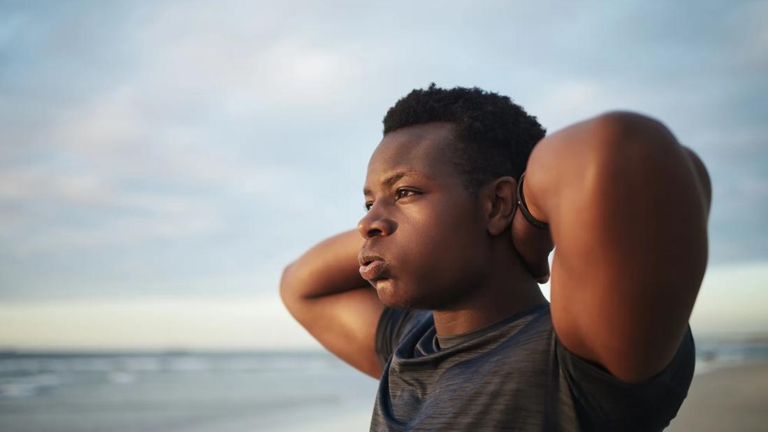 Male standing on beach with hands behind his head, staring into distance and exhaling