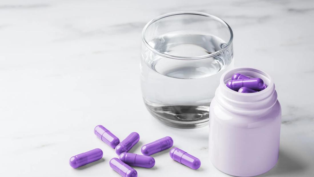 Glass of water on table, with medicine bottle of purple capsules, and capsules on table