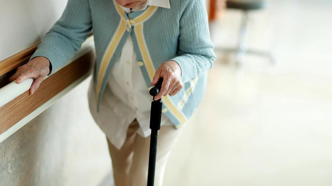 Elderly woman using a cane and handrail on wall walking down a hall