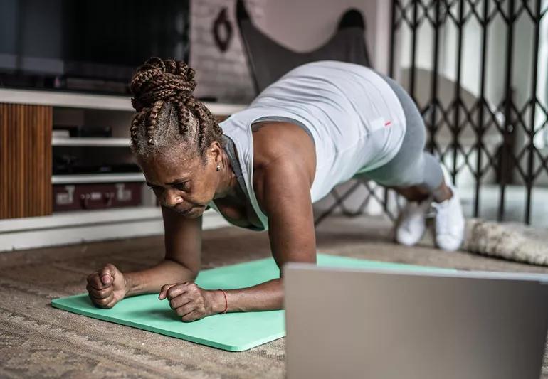 Elder woman performs a plank on a green mat on the floor while in an online exercise class.