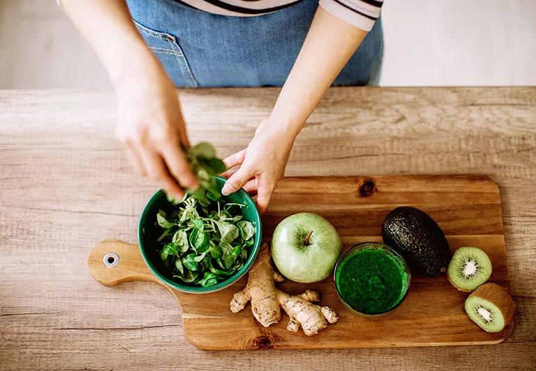 A person fills a bowl with arugula next to a cutting board with ginger, an apple, cilantro, avocado and a kiwi.