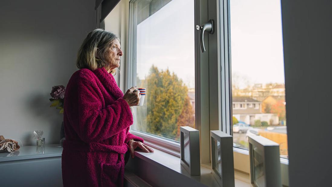 Older woman in bathrobe, holding coffee cup, looking window into the neighborhood