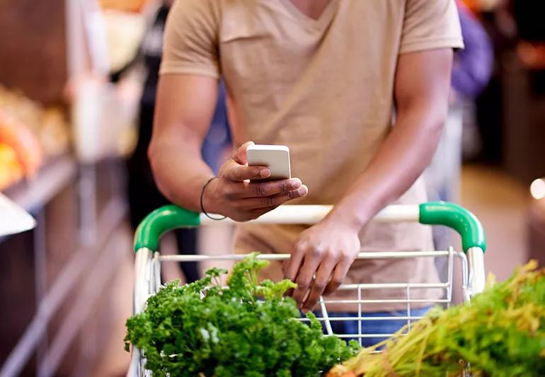 person pushing shopping cart with greens while looking at phone