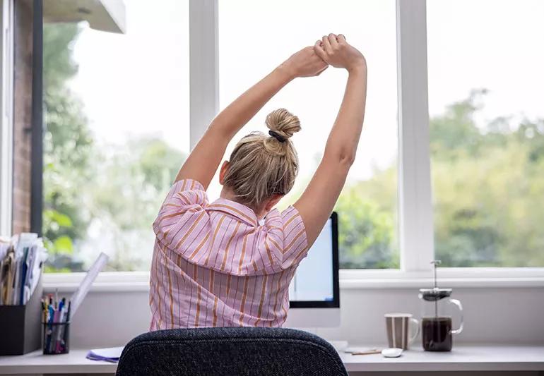 A person at their work desk holding their hands together above their head and stretching to the side