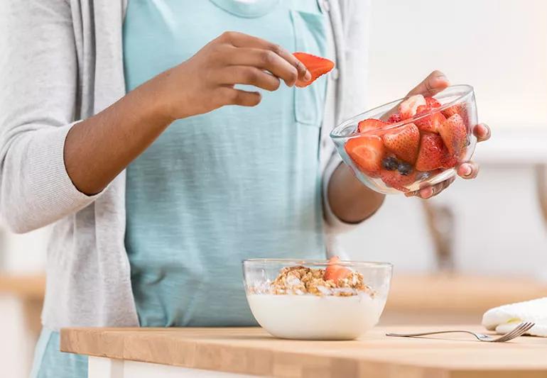 A woman is putting strawberries and blueberries into a bowl of cereal.