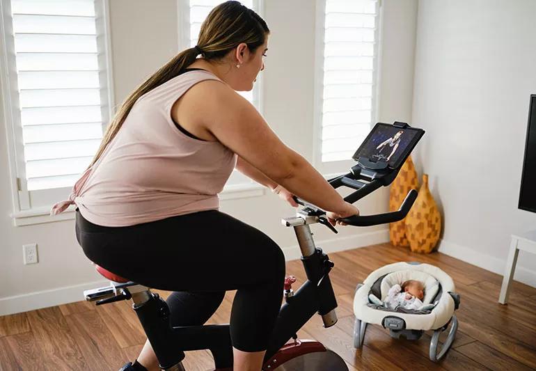 Woman riding an exercise bike while a baby sleeps nearby