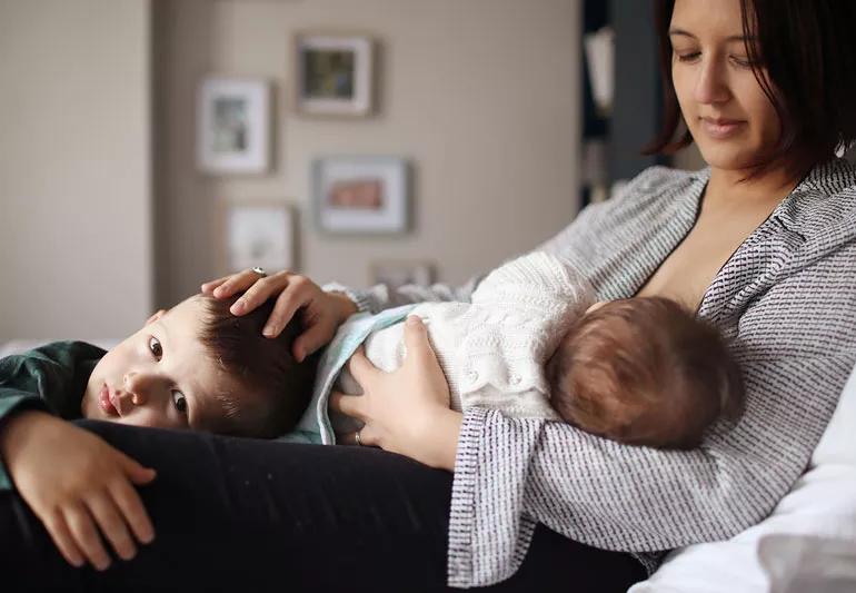 Woman breastfeeding baby on couch with younger sibling present.