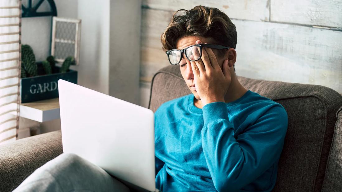 Person wearing glasses sitting on couch holding laptop, rubbing eyes, looking tired