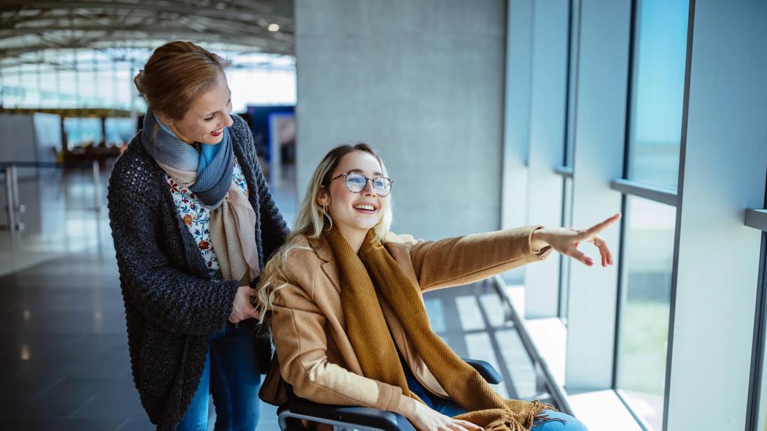 Smiling caretaker pushing smiling person in wheelchair at the airport, looking out the window